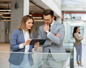 Ein Mann und eine Frau in formeller Kleidung betrachten gemeinsam ein Tablet in einem modernen Büro. Im Hintergrund sind weitere Personen zu sehen.