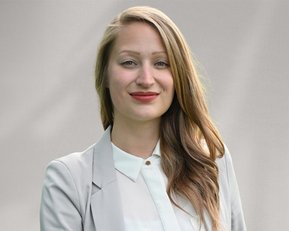 Eine lächelnde Frau mit langen, lockigen Haaren trägt einen hellen Blazer und ein Hemd. Der Hintergrund ist neutral und unauffällig.