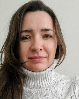 Eine Frau mit langen, braunen Haaren trägt einen hellen, gestrickten Rollkragenpullover. Sie schaut direkt in die Kamera.