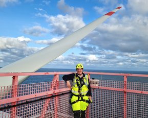 Eine Person in Sicherheitsausrüstung steht auf einer Plattform neben einem Windradblatt, mit Blick auf das Meer und den Himmel.