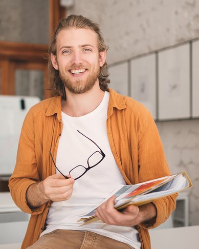 Ein junger Mann mit langen Haaren sitzt auf einem Tisch, hält eine Mappe mit Unterlagen und eine Brille in der Hand. Er trägt ein orangefarbenes Hemd und lächelt.