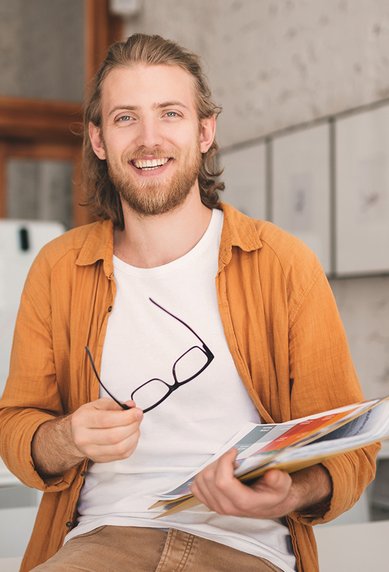 Ein junger Mann mit langen Haaren sitzt auf einem Tisch, hält eine Mappe mit Unterlagen und eine Brille in der Hand. Er trägt ein orangefarbenes Hemd und lächelt.
