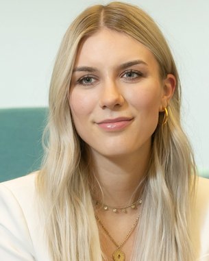 Eine junge Frau mit langen, blonden Haaren und einem weißen Blazer sitzt vor einem grünen Stuhl. Sie hat eine neutrale Miene und trägt mehrere Schmuckstücke.
