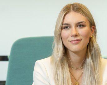 Eine junge Frau mit langen, blonden Haaren und einem weißen Blazer sitzt vor einem grünen Stuhl. Sie hat eine neutrale Miene und trägt mehrere Schmuckstücke.