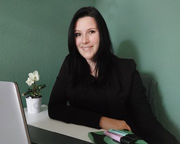 Eine Frau mit langen, dunklen Haaren sitzt an einem Tisch, lächelt und trägt einen schwarzen Blazer. Auf dem Tisch befinden sich ein Laptop, Schreibutensilien und eine kleine Pflanze in einem Topf.