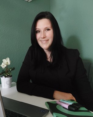 Eine Frau mit langen, dunklen Haaren sitzt an einem Tisch, lächelt und trägt einen schwarzen Blazer. Auf dem Tisch befinden sich ein Laptop, Schreibutensilien und eine kleine Pflanze in einem Topf.