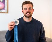Ein junger Mann hält eine Medaille mit blauer Schleife in der Hand. Er trägt einen dunkelblauen Pullover und sitzt in einem hellen Raum.