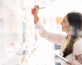 Zwei Frauen arbeiten gemeinsam an einer Wand mit Haftnotizen. Eine Frau schreibt mit einem Stift auf eine Haftnotiz, während die andere sie beobachtet und Notizen macht.
