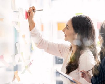 Zwei Frauen arbeiten gemeinsam an einer Wand mit Haftnotizen. Eine Frau schreibt mit einem Stift auf eine Haftnotiz, während die andere sie beobachtet und Notizen macht.