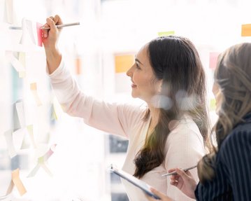 Zwei Frauen arbeiten gemeinsam an einer Wand mit Haftnotizen. Eine Frau schreibt mit einem Stift auf eine Haftnotiz, während die andere sie beobachtet und Notizen macht.