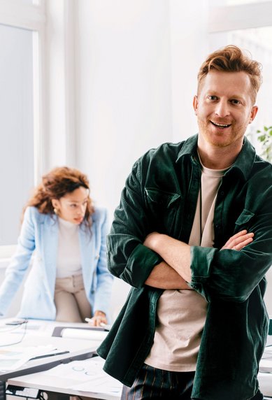 Ein Mann mit verschränkten Armen steht in einem modernen Büro, während drei Kollegen an Schreibtischen arbeiten.