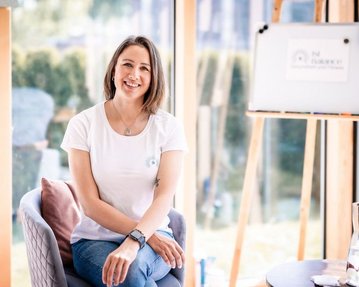 Eine lächelnde Frau sitzt auf einem Stuhl, trägt ein weißes T-Shirt und Jeans, umgeben von großen Fenstern und einem Flipchart im Hintergrund.