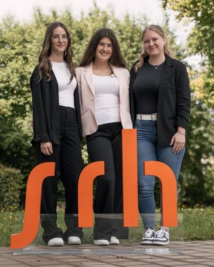 Drei Frauen stehen vor einem großen, orangefarbenen Logo mit den Buchstaben "sdh" in einem Park. Sie tragen formelle Kleidung.