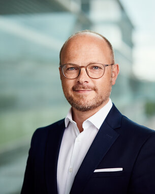 Mann mit Brille und Anzug steht vor einer Glaswand, lächelt leicht und schaut in die Kamera. Heller Hintergrund mit unscharfen Gebäuden.