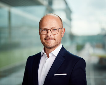 Mann mit Brille und Anzug steht vor einer Glaswand, lächelt leicht und schaut in die Kamera. Heller Hintergrund mit unscharfen Gebäuden.