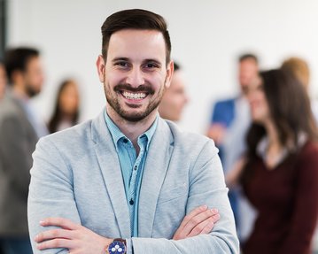 Fernstudium Master Wirtschaftspsychologie & Leadership | SRH Fernhochschule Ein Mann im Anzug mit einem breiten Lächeln und gekreuzten Armen steht im Vordergrund, während eine Gruppe von Menschen im Hintergrund miteinander spricht.