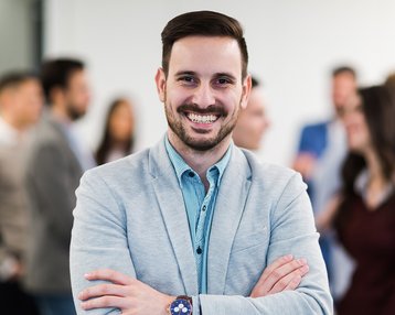 Ein Mann im Anzug mit einem breiten Lächeln und gekreuzten Armen steht im Vordergrund, während eine Gruppe von Menschen im Hintergrund miteinander spricht.