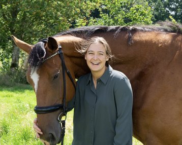 Eine Person steht neben einem braunen Pferd und lächelt. Die Person trägt ein graues Hemd und hält das Pferd am Halfter. Im Hintergrund sind Bäume und eine grüne Wiese zu sehen.