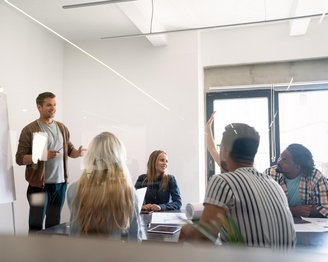 Ein Mann präsentiert in einem Besprechungsraum, während mehrere Personen aufmerksam zuhören und Fragen stellen. An einer Wand steht ein Flipchart mit Diagrammen.