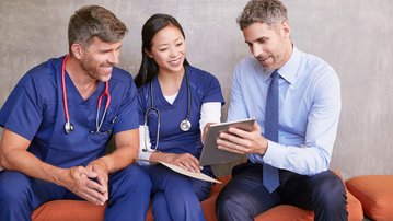 Drei Personen in medizinischer Kleidung und Anzug sitzen auf einer Couch und schauen auf ein Tablet. Zwei Männer und eine Frau lächeln, während sie Informationen auf dem Gerät besprechen.