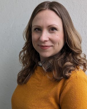 Eine Frau mit lockigem, braunem Haar trägt einen orangefarbenen Pullover und schaut zur Kamera. Der Hintergrund ist hell und neutral.