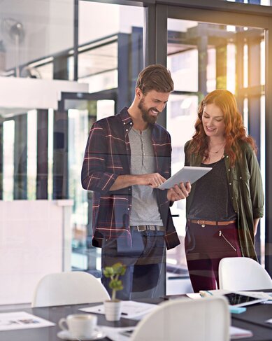Zwei Personen stehen in einem modernen Büro und betrachten gemeinsam ein Tablet. Der Mann trägt ein kariertes Hemd, die Frau eine Jacke. Auf dem Tisch sind Unterlagen und Tassen sichtbar.