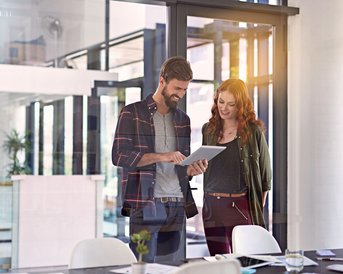 Psychologie in Organisationen | SRH Fernhochschule Zwei Personen stehen in einem modernen Büro und betrachten gemeinsam ein Tablet. Der Mann trägt ein kariertes Hemd, die Frau eine Jacke. Auf dem Tisch sind Unterlagen und Tassen sichtbar.