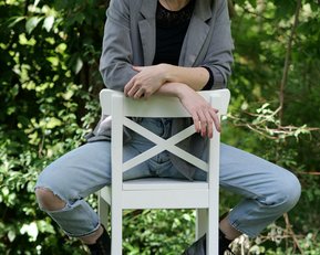 Eine Frau sitzt lässig auf einem weißen Hocker, trägt eine graue Jacke und Jeans, umgeben von grünen Pflanzen.