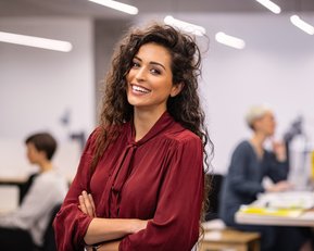 Eine Frau mit lockigem Haar und rotem Oberteil lächelt in einem modernen Büro. Im Hintergrund sind weitere Personen bei der Arbeit zu sehen.