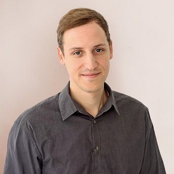 Ein junger Mann mit kurzen, braunen Haaren trägt ein graues Hemd und steht vor einer hellen Wand. Er lächelt leicht und schaut direkt in die Kamera.