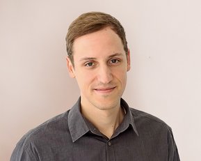 Ein junger Mann mit kurzen, braunen Haaren trägt ein graues Hemd und steht vor einer hellen Wand. Er lächelt leicht und schaut direkt in die Kamera.