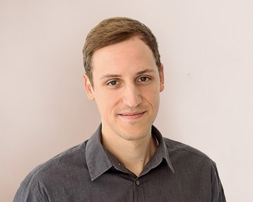 Ein junger Mann mit kurzen, braunen Haaren trägt ein graues Hemd und steht vor einer hellen Wand. Er lächelt leicht und schaut direkt in die Kamera.