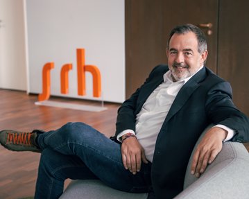 Ein Mann sitzt entspannt auf einem grauen Sofa, trägt einen schwarzen Blazer und Jeans, mit orangefarbenen Schuhen. Im Hintergrund steht das orangefarbene Logo "srh".