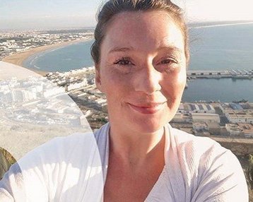 Frau mit lächelndem Gesicht, die ein Selfie vor einer Küstenlandschaft macht. Im Hintergrund sind das Meer und eine Stadtansicht mit Hafenanlagen sichtbar.