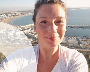 Frau mit lächelndem Gesicht, die ein Selfie vor einer Küstenlandschaft macht. Im Hintergrund sind das Meer und eine Stadtansicht mit Hafenanlagen sichtbar.