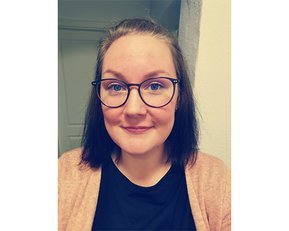 Eine Person mit kurzen, dunklen Haaren und Brille lächelt in die Kamera. Sie trägt ein schwarzes T-Shirt und einen hellen Cardigan. Der Hintergrund zeigt eine Wand und eine Tür.
