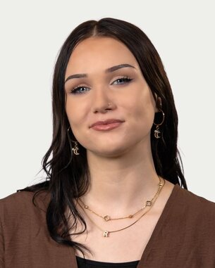 Junge Frau mit langen, dunklen Haaren, trägt goldene Ohrringe und Halsketten, schaut direkt in die Kamera.
