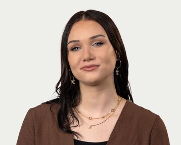 Junge Frau mit langen, dunklen Haaren, trägt goldene Ohrringe und Halsketten, schaut direkt in die Kamera.