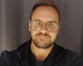 Ein Mann mit Brille und Bart lächelt in die Kamera. Er trägt eine dunkle Jacke und ein T-Shirt. Der Hintergrund ist grau.
