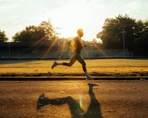 Ein Läufer sprintet auf einer Straße, während die Sonne im Hintergrund aufgeht und lange Schatten wirft.
