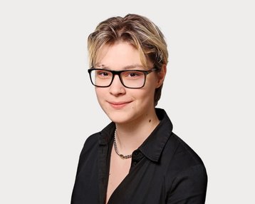 Eine Person mit kurzen, blonden Haaren und Brille trägt ein schwarzes Hemd. Der Hintergrund ist neutral und hell.