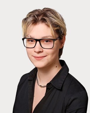 Eine Person mit kurzen, blonden Haaren und Brille trägt ein schwarzes Hemd. Der Hintergrund ist neutral und hell.