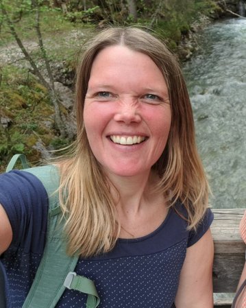 Eine Frau mit langen, blonden Haaren lächelt in die Kamera, während sie an einem Flussufer steht, umgeben von Bäumen.