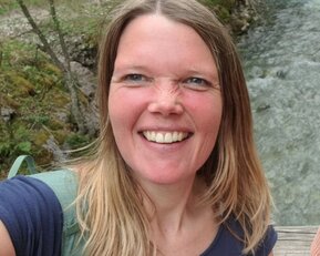 Eine Frau mit langen, blonden Haaren lächelt in die Kamera, während sie an einem Flussufer steht, umgeben von Bäumen.