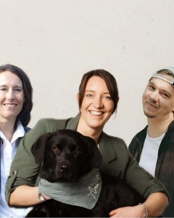 Drei Personen stehen zusammen, eine hält einen schwarzen Labrador, während die anderen lächeln. Alle tragen legere Kleidung.