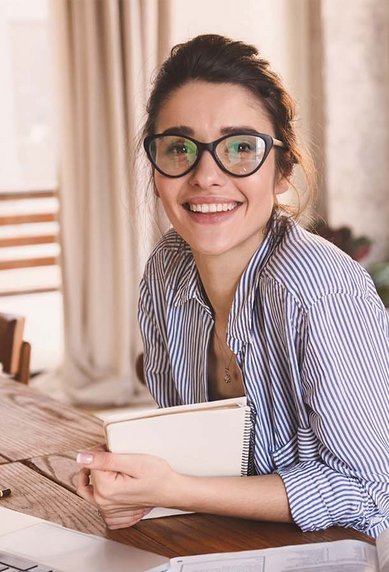 Eine lächelnde Frau mit Brille sitzt an einem Tisch, umgeben von Pflanzen und einem Laptop. Sie hält ein Notizbuch in der Hand und trägt ein gestreiftes Hemd.