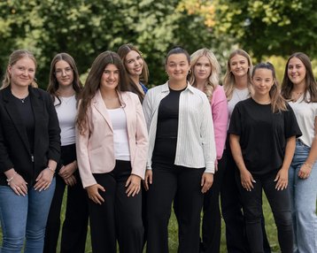 Eine Gruppe von neun Frauen steht zusammen im Freien, lächelnd und in formeller Kleidung, umgeben von grüner Landschaft.