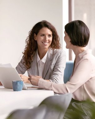 Zwei Frauen sitzen an einem Tisch, eine von ihnen lächelt und hält ein Dokument, während die andere aufmerksam zuhört. Ein Laptop und eine Tasse stehen auf dem Tisch.