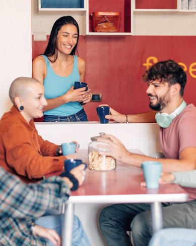 Eine Gruppe junger Menschen sitzt entspannt an einem Tisch und unterhält sich bei Kaffee oder Tee. Eine weitere Person steht lächelnd hinter einer Theke mit der Aufschrift „free food“. Die Atmosphäre wirkt freundlich und einladend, wie in einem Gemeinschaftsraum oder sozialen Treffpunkt.