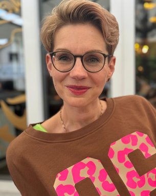 Frau mit kurzen, blonden Haaren und Brille trägt einen braunen Pullover mit großen, pinken Zahlen. Sie lächelt und steht vor einem hellen Hintergrund mit unscharfen Details.